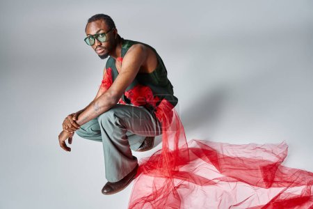 appealing african american man in fashionable attire squatting on gray backdrop, fashion concept