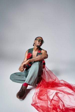 atractivo hombre afroamericano con tela de tul rojo en su chaleco sentado en el suelo, concepto de moda