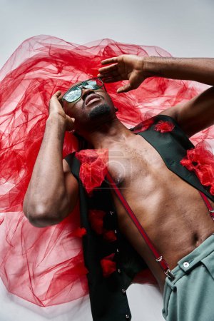Téléchargez les photos : Attrayant homme afro-américain à la mode avec des lunettes de soleil en tenue vibrante couché sur le sol, la mode - en image libre de droit