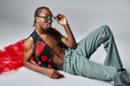 Téléchargez les photos : Beau jeune homme afro-américain en tenue vibrante avec tissu en tulle rouge inclinable sur le sol - en image libre de droit