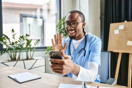 Photo for Merry african american doctor with glasses waving at mobile phone camera and smiling happily - Royalty Free Image