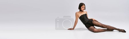 Photo for Pretty asian woman with short hair sitting in black corset and polka dot tights on white floor - Royalty Free Image