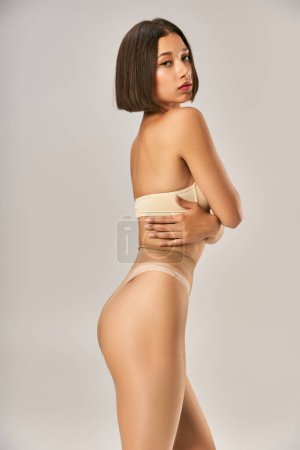 atractiva y joven mujer asiática en pantimedias sin costuras y ropa interior beige posando sobre fondo gris