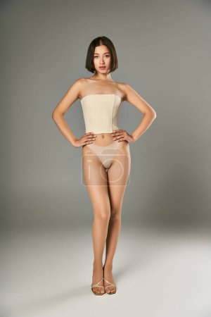 young asian woman in fishnet tights and panties posing with hands on hips on grey background