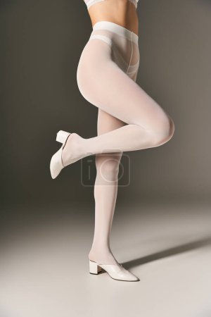 geschnittenes Foto einer schlanken Frau in hauchdünnen weißen Strumpfhosen und High Heels, die auf grauem Hintergrund posiert