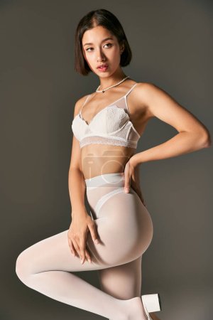 Foto de Hermosa mujer asiática en blanco sujetador de encaje y pantimedias posando con la mano en la cadera sobre fondo gris - Imagen libre de derechos