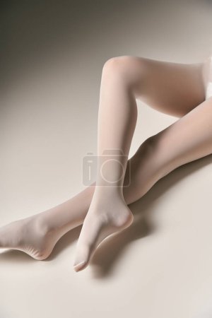 cropped view of young woman in sheer white trendy pantyhose sitting on grey background, fashion