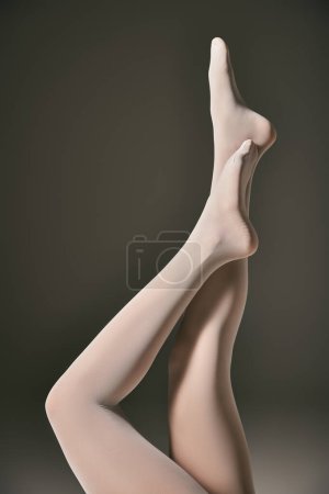 vista recortada de modelo joven en pantimedias blanco puro posando con las piernas levantadas sobre fondo gris oscuro