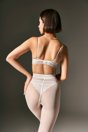 back view of young woman in white underwear and pantyhose posing with hand on hip on grey background