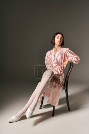 Photo for Alluring young asian woman in pink silk robe and white tights sitting on chair on grey background - Royalty Free Image