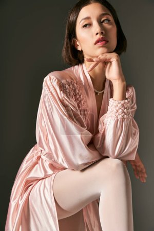 femme asiatique rêveuse et jeune en robe de soie rose et collants blancs posant sur fond gris