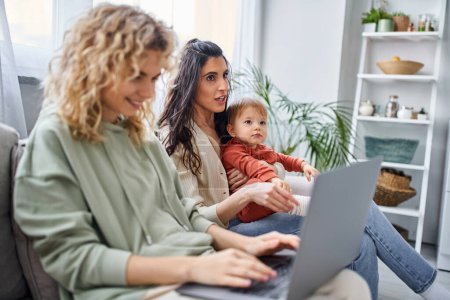 Téléchargez les photos : Se concentrer sur la femme brune avec bébé fille assis sur le canapé près de son partenaire flou avec ordinateur portable, famille - en image libre de droit