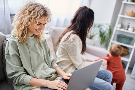 focus on blonde woman with laptop sitting on sofa near her blurred partner with their baby, family