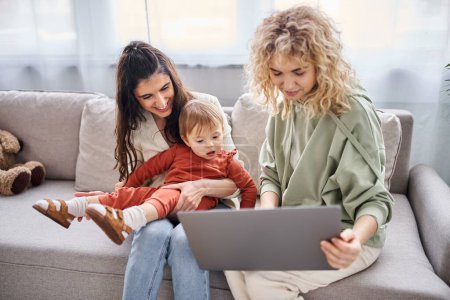 Téléchargez les photos : Joyeux amour couple lesbien assis sur canapé avec leur bébé fille regardant ordinateur portable, concept de famille - en image libre de droit