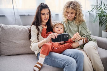 Téléchargez les photos : Attrayant couple lgbt assis sur le canapé avec leur petite fille regardant le téléphone mobile, concept de famille - en image libre de droit
