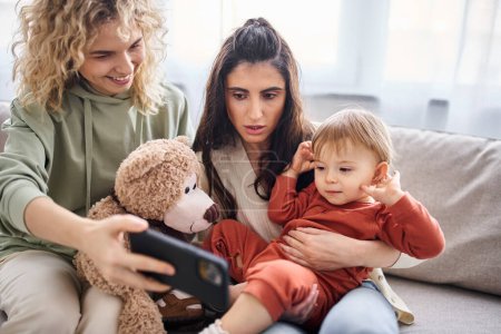 caring lgbt couple on sofa with their baby girl looking at phone holding teddy bear, family concept