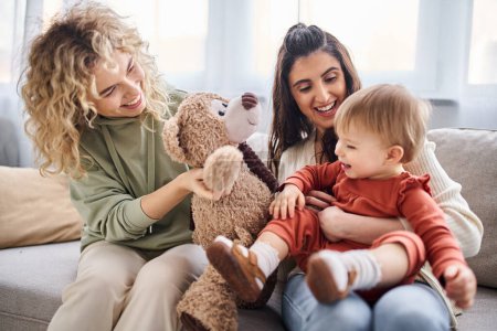 Téléchargez les photos : Beau couple lesbien joyeux avec leur fille tout-petit et ours en peluche sur canapé, concept de famille - en image libre de droit