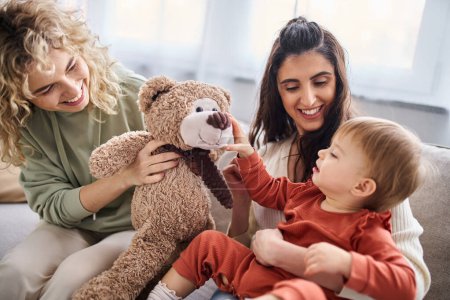 Téléchargez les photos : Beau couple de lesbiennes heureux avec leur fille tout-petit et ours en peluche sur le canapé, concept de famille - en image libre de droit