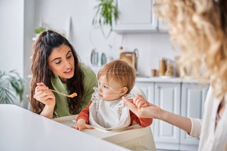Téléchargez les photos : Focus sur femme brune joyeuse avec petite fille prenant le petit déjeuner à côté de partenaire flou, famille - en image libre de droit