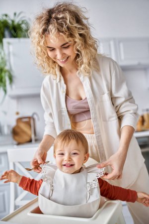 Photo for Joyous beautiful blonde woman putting bib on her pretty jolly baby girl before breakfast, family - Royalty Free Image