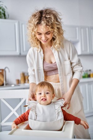 Photo for Attractive woman in homewear putting bib on her joyous baby girl before breakfast, family concept - Royalty Free Image
