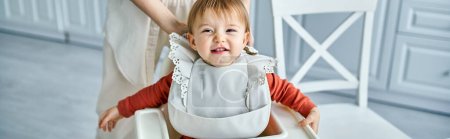 Photo for Caring woman putting bib on her pretty cheerful baby girl before breakfast, family concept, banner - Royalty Free Image