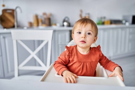 Charmantes Kleinkind in orangefarbenem Pullover sitzt beim Frühstück auf dem Hochstuhl und blickt in die Kamera