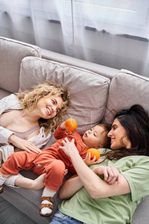 Téléchargez les photos : Joyeux couple lgbt couché sur le canapé avec leur jolie fille tenant des mandarines, concept de famille - en image libre de droit