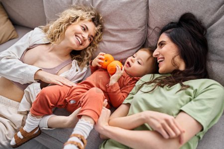 Téléchargez les photos : Heureux couple lgbt couché sur canapé avec leur jolie fille tenant des mandarines, concept de famille - en image libre de droit