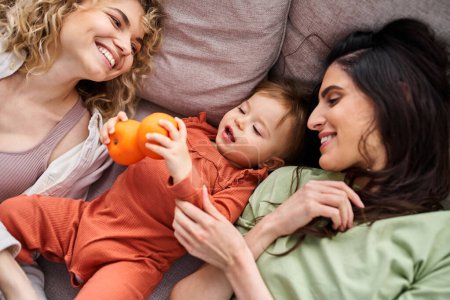 Téléchargez les photos : Beau couple joyeux lgbt couché sur le canapé avec leur bébé fille tenant des mandarines, concept de famille - en image libre de droit