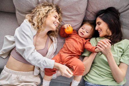 Téléchargez les photos : Beau couple joyeux lgbt couché sur le canapé avec leur petite fille tenant des mandarines, concept de famille - en image libre de droit