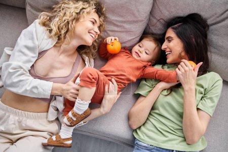 Téléchargez les photos : Attrayant couple joyeux lgbt couché sur le canapé avec leur bébé fille tenant des mandarines, concept de famille - en image libre de droit