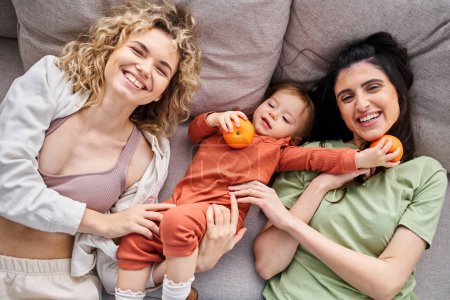 Téléchargez les photos : Attrayant couple gai lgbt couché sur le canapé avec leur bébé fille tenant des mandarines, concept de famille - en image libre de droit