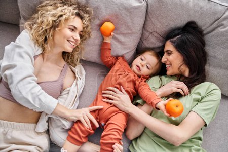 Téléchargez les photos : Amour joyeux couple lgbt couché sur le canapé avec leur bébé fille tenant des mandarines, concept de famille - en image libre de droit
