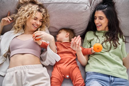 Téléchargez les photos : Amour heureux couple lgbt couché sur le canapé avec leur bébé fille tenant des mandarines, concept de famille - en image libre de droit