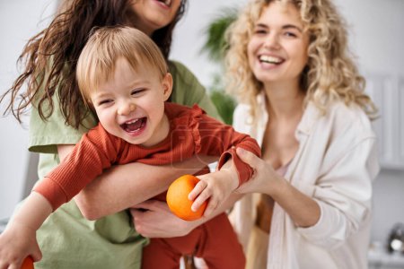 Téléchargez les photos : Joyeux couple lesbien s'amuser avec leur mignon bébé fille tenant des mandarines, famille - en image libre de droit