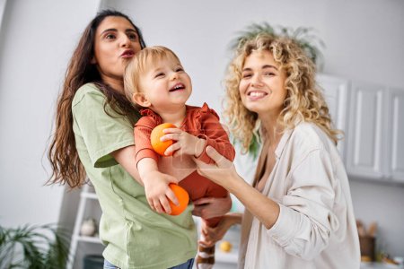 Téléchargez les photos : Joyeux joli couple lgbt s'amuser avec leur jolie petite fille tenant des mandarines, famille - en image libre de droit