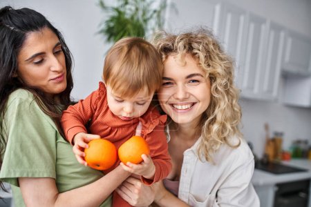 Téléchargez les photos : Joyeux joli couple lgbt s'amuser avec leur jolie petite fille tenant des mandarines, famille - en image libre de droit