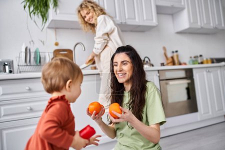 Téléchargez les photos : Se concentrer sur la femme joyeuse avec sa fille tenant des mandarines avec son partenaire blond flou sur fond - en image libre de droit