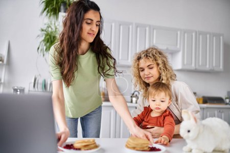 Téléchargez les photos : Beau couple lesbien joyeux ayant pancakes sur le petit déjeuner avec leur petite fille, concept de famille - en image libre de droit