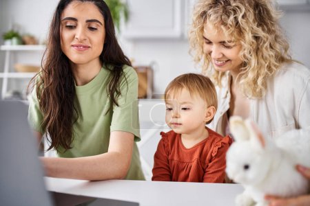 beautiful lesbian couple in homewear watching movies with their baby girl on laptop, family concept