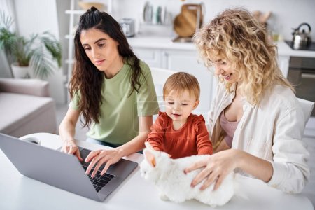 positives lesbisches Paar in Homewear, das mit seinem kleinen Mädchen auf dem Laptop Filme guckt, Familienkonzept
