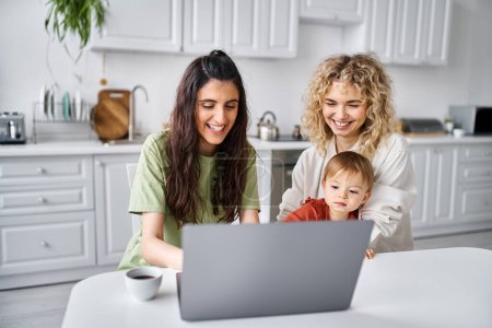 Téléchargez les photos : Heureux couple lgbt regarder des films avec leur fille sur ordinateur portable sur la cuisine, parentalité moderne - en image libre de droit