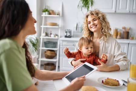 bonded lgbt couple having breakfast with their baby girl and looking at tablet, family concept
