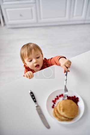 adorable niña en cómodo suéter naranja tratando de llegar a un poco de desayuno en la mesa