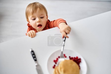 Foto de Adorable niña en suéter naranja acogedor tratando de llegar a un poco de desayuno en la mesa - Imagen libre de derechos