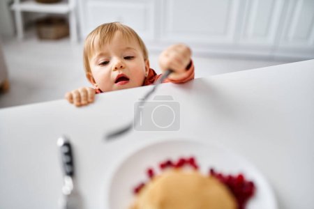 jolie petite fille en pull orange confortable essayant de rejoindre un petit déjeuner sur la table