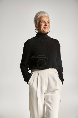 retrato de mujer de negocios de mediana edad feliz en traje elegante posando con las manos en bolsillos en gris