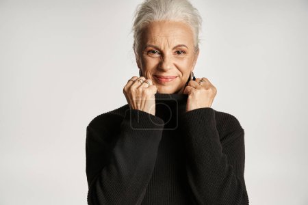 Téléchargez les photos : Portrait de femme d'affaires d'âge moyen ajustant le col du pull à col roulé sur fond gris - en image libre de droit