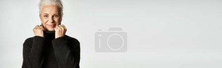Téléchargez les photos : Portrait de femme d'affaires d'âge moyen ajustant le col du pull à col roulé sur fond gris - en image libre de droit